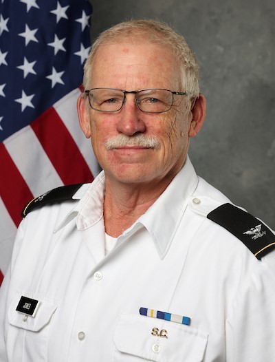 Dr. Wesley M. Jones Jr., a man with glasses, gray hair, and a mustache, wears a white military uniform as he stands before an American flag and gray background.