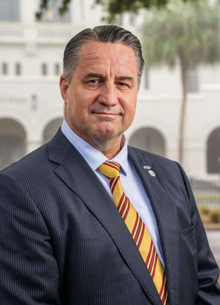 Bill Connor wearing a dark suit and yellow striped tie, standing outdoors at The Citadel.