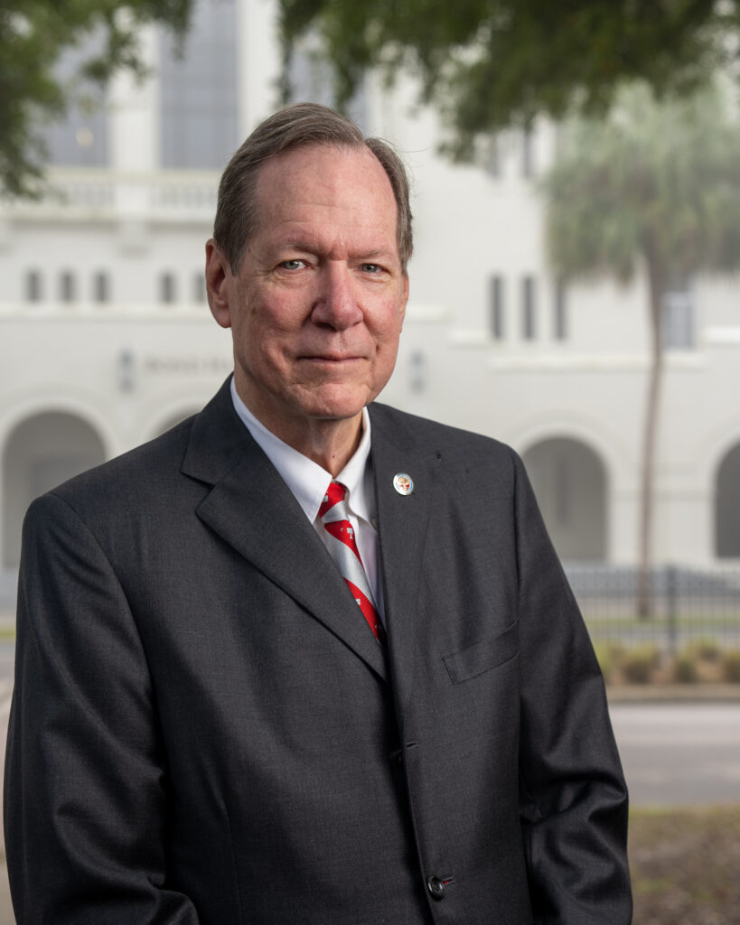 Greg Delleney wearing a dark suit and red striped tie, standing outdoors at The Citadel.