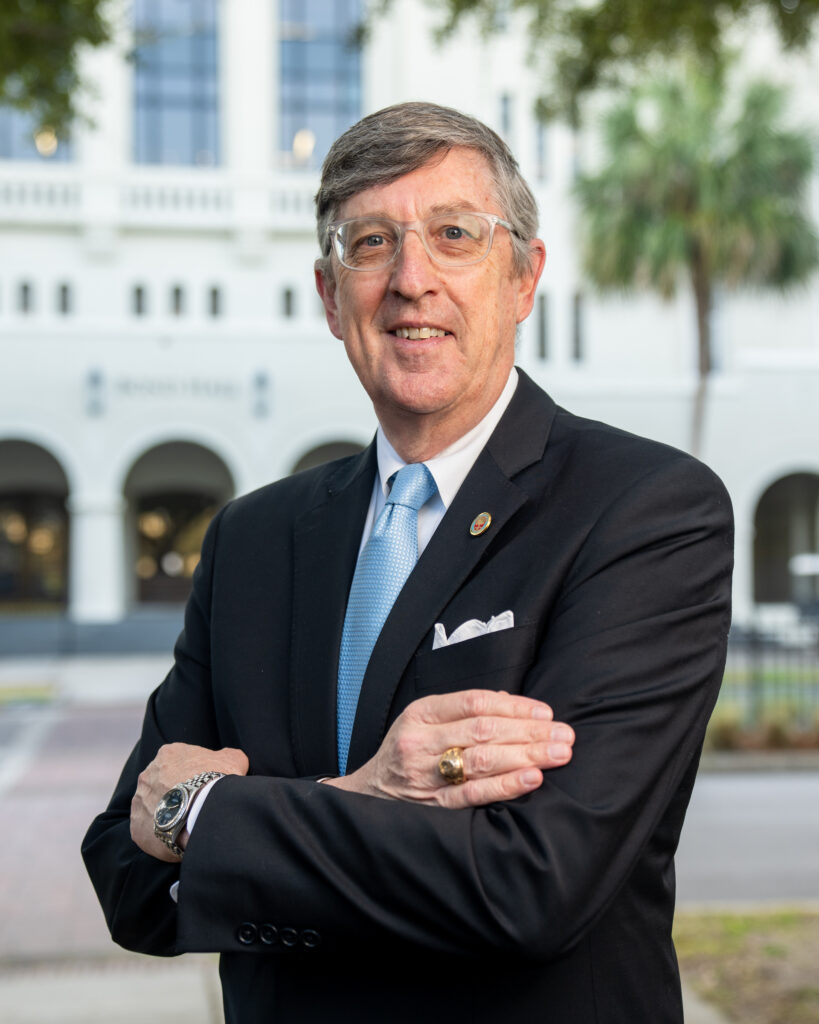 Professional headshot of Douglas Snyder in a black suit and blue tie standing outdoors in front of a white building with arches at The Citadel.