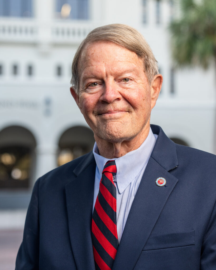Professional headshot of Gene Pinson in a navy suit and red striped tie standing outdoors at The Citadel.
