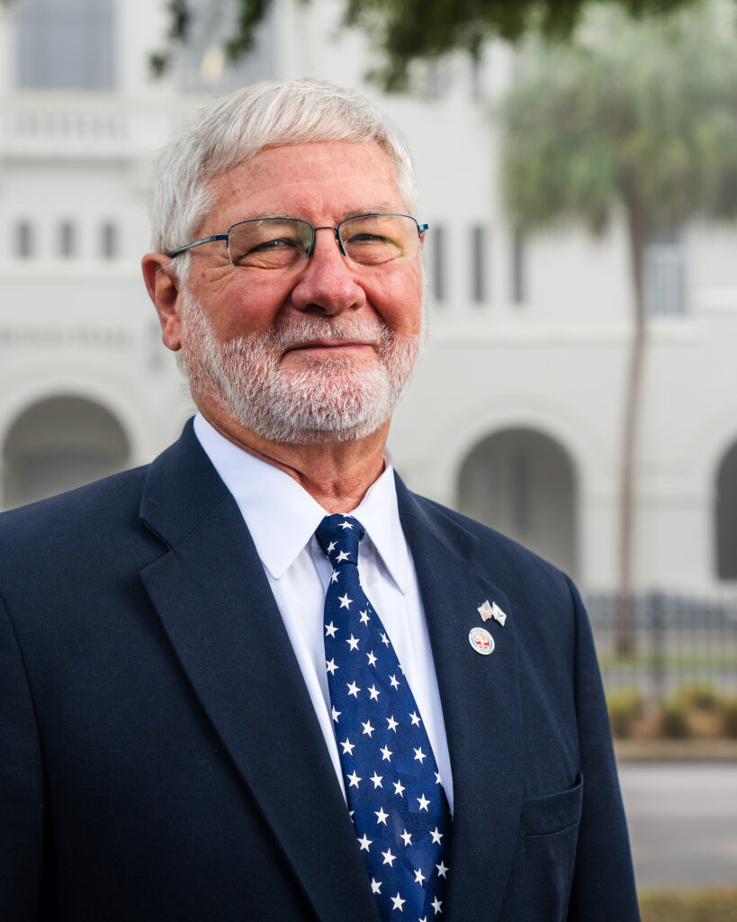 Professional headshot of John Dominick in a black suit and blue star tie standing outdoors at The Citadel.