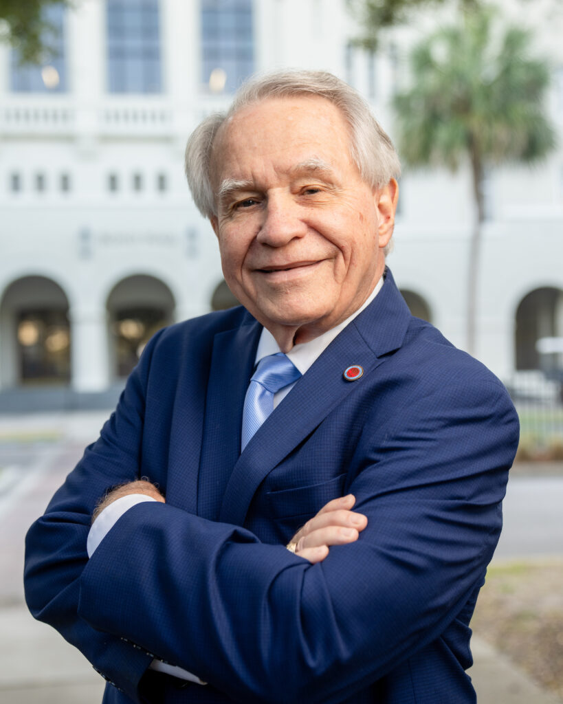 Professional headshot of William Jenkinson in a navy suit and blue striped tie standing outdoors in front of a white building with arches and a palm tree at The Citadel.