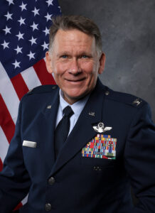 Dr. Michael R. Weeks, in a U.S. Air Force dress uniform, stands smiling before an American flag, proudly displaying medals and insignia on his jacket.