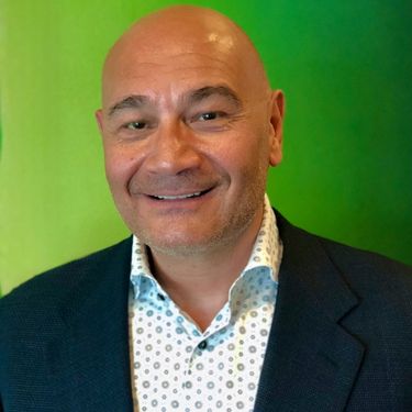 Headshot of Mark Dajani smiling, wearing a dark blazer and patterned shirt against a green background.