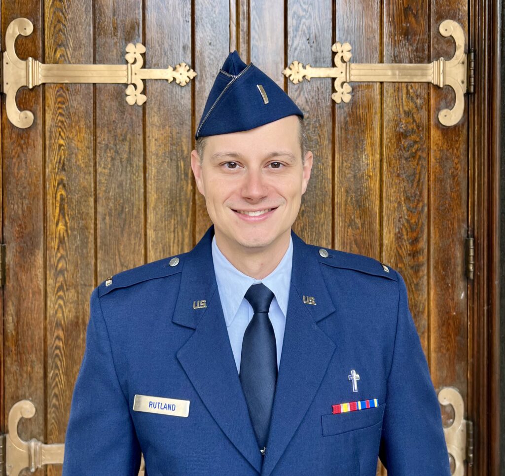 A person in a blue military uniform and cap stands smiling in front of large wooden doors. The name tag reads Associate Chaplain Stephen Rutland, member of the Chapel Team at The Citadel Faith Specific Ministries.