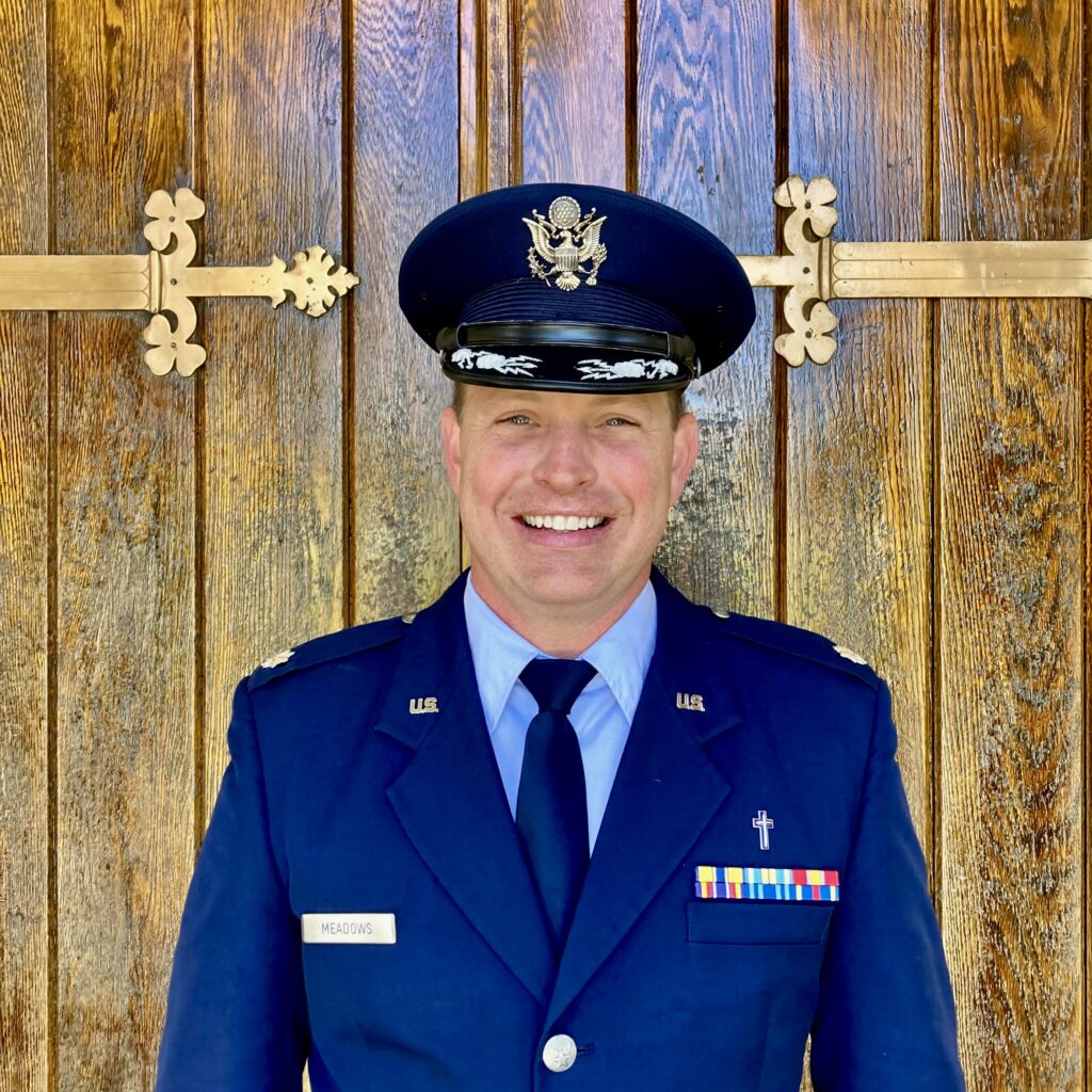 A smiling man in a blue U.S. Air Force dress uniform and cap stands before large wooden doors with brass hinges, representing associate chaplain Stephen Rutland of The Citadel Faith Specific Ministries.