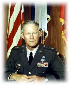 Portrait of a U.S. Army general in dress uniform seated in front of American and military flags.
