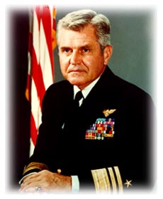 Portrait of a U.S. Navy admiral in dress uniform standing in front of an American flag.