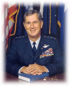 Portrait of a U.S. Air Force officer in dress uniform seated at a desk with military flags behind him.