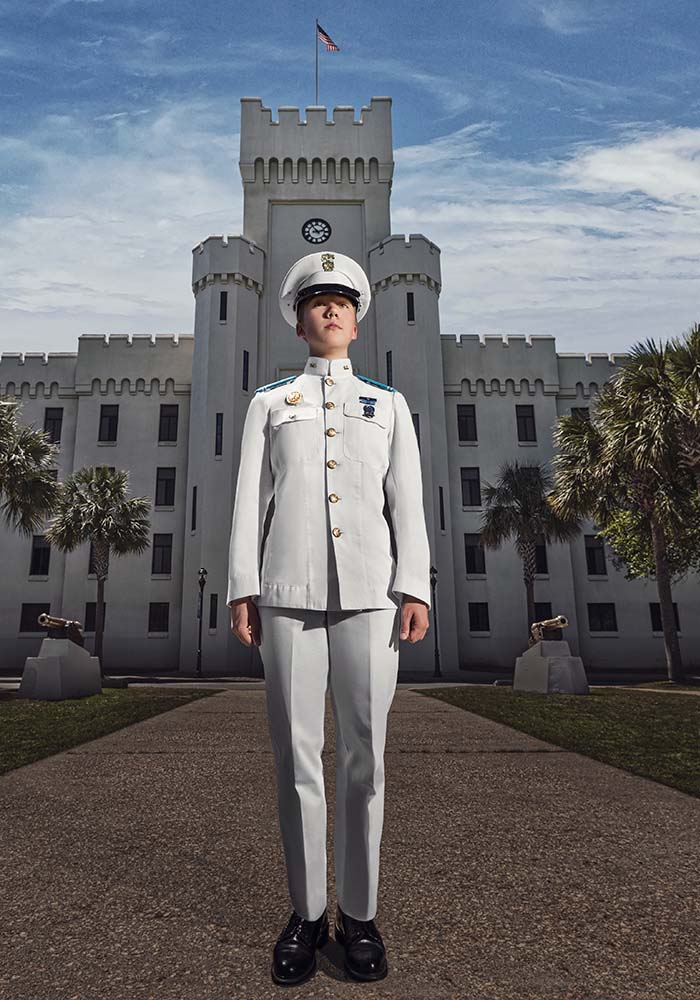 A photo of a cadet in front of a barrack in full white uniform. 