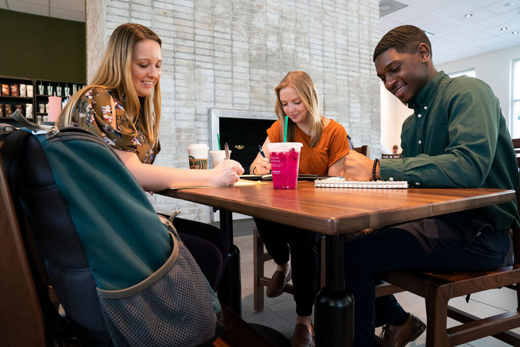 Three students studying together at a table with notebooks and drinks.