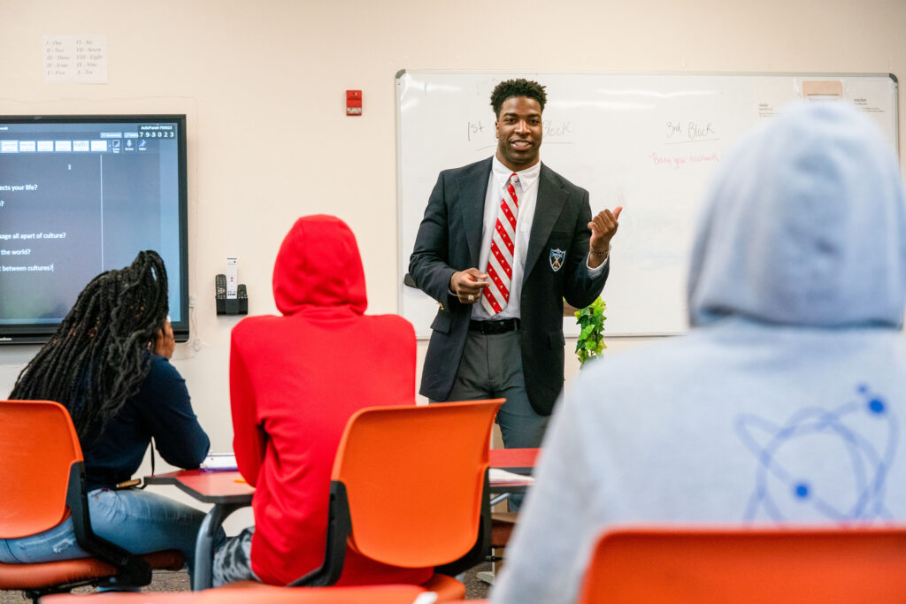 A Citadel education major leads a classroom discussion with students seated around tables during a lesson.
