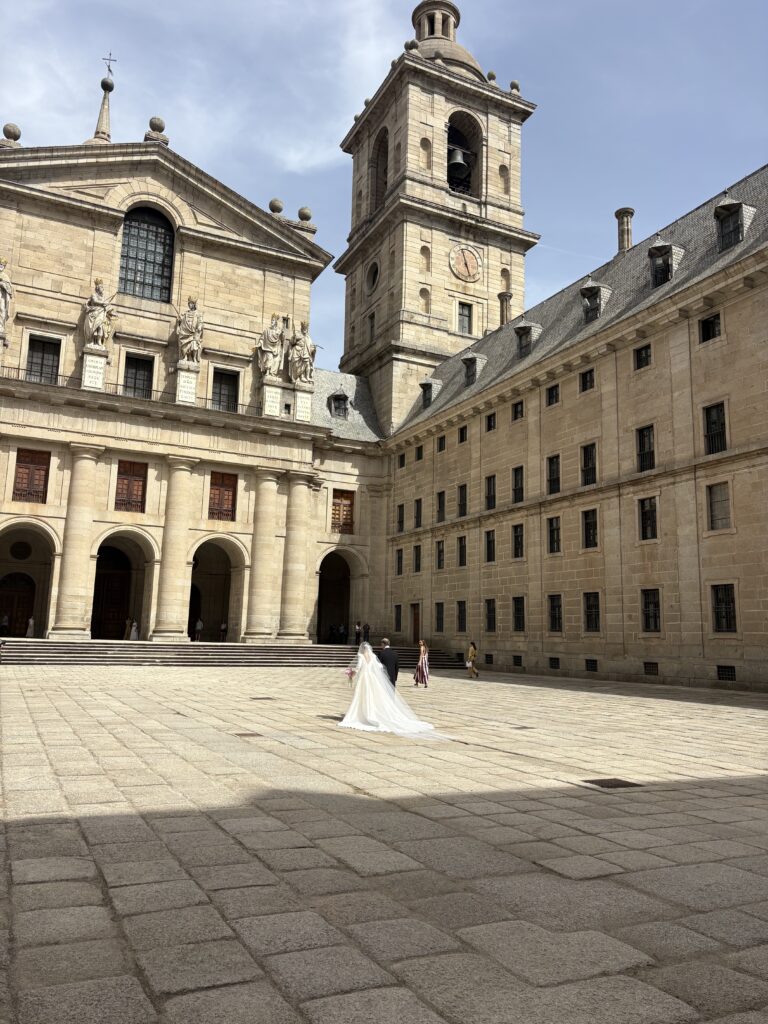Bride and groom walking across a sunlit stone courtyard surrounded by historic buildings and a bell tower.