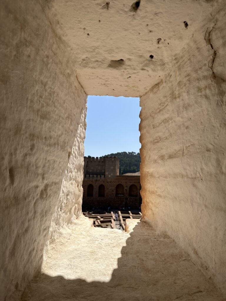 View through a narrow stone opening toward a historic fortress courtyard under a clear blue sky.