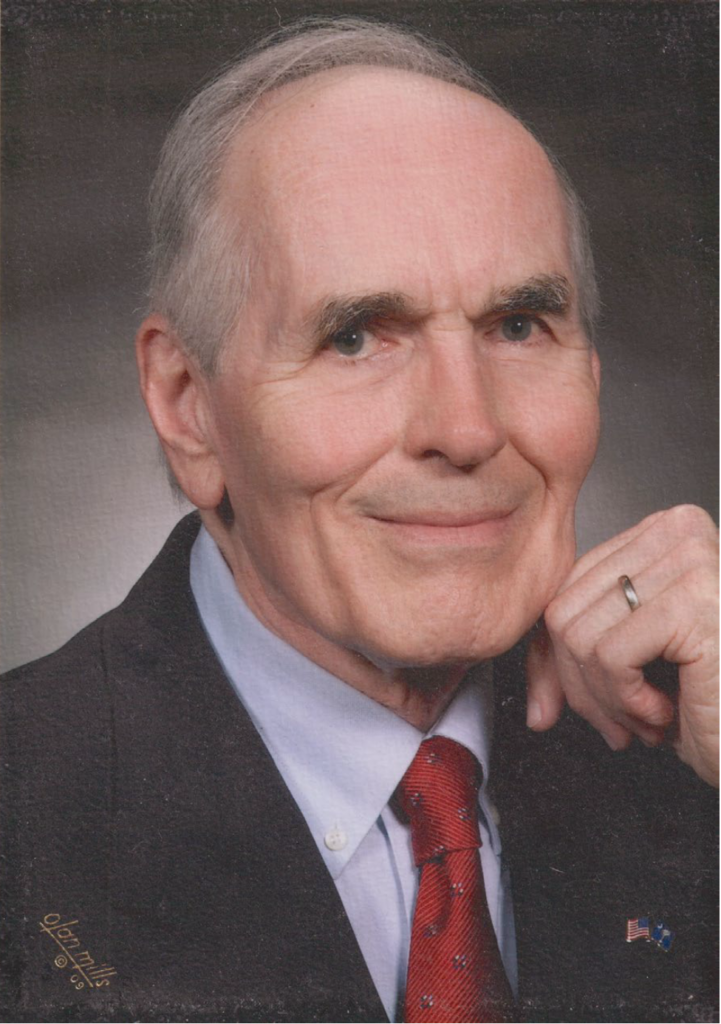 Portrait of Charles Watson Jr. in a dark suit and red tie, resting his hand near his chin and smiling at the camera.