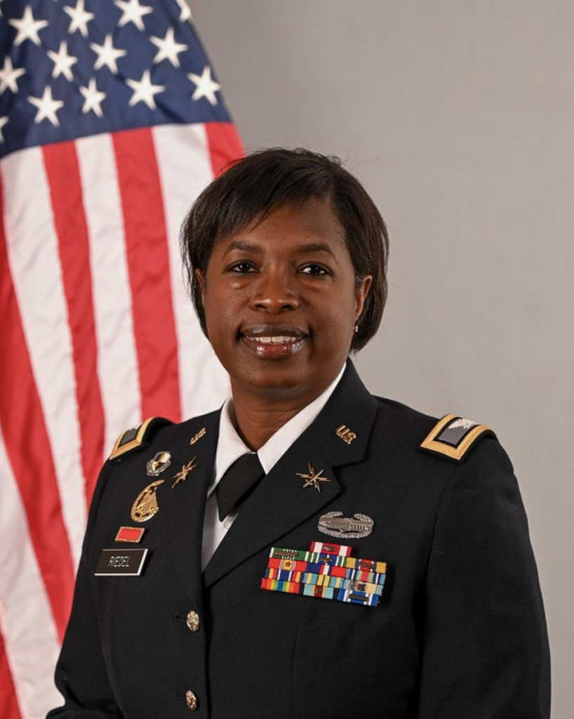 Portrait of Col Linda Riedel in a U.S. Army uniform with medals, standing in front of an American flag.