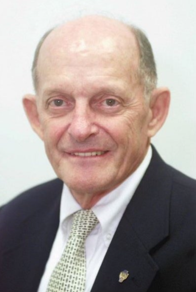 Portrait of an Col. Ned Sanders in a suit and patterned tie, smiling at the camera against a plain background.
