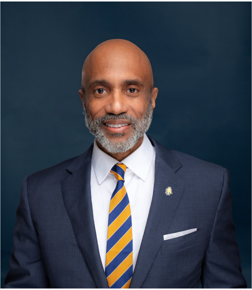 Portrait of James Martin, wearing a navy suit and blue-and-gold striped tie, smiling against a dark blue background