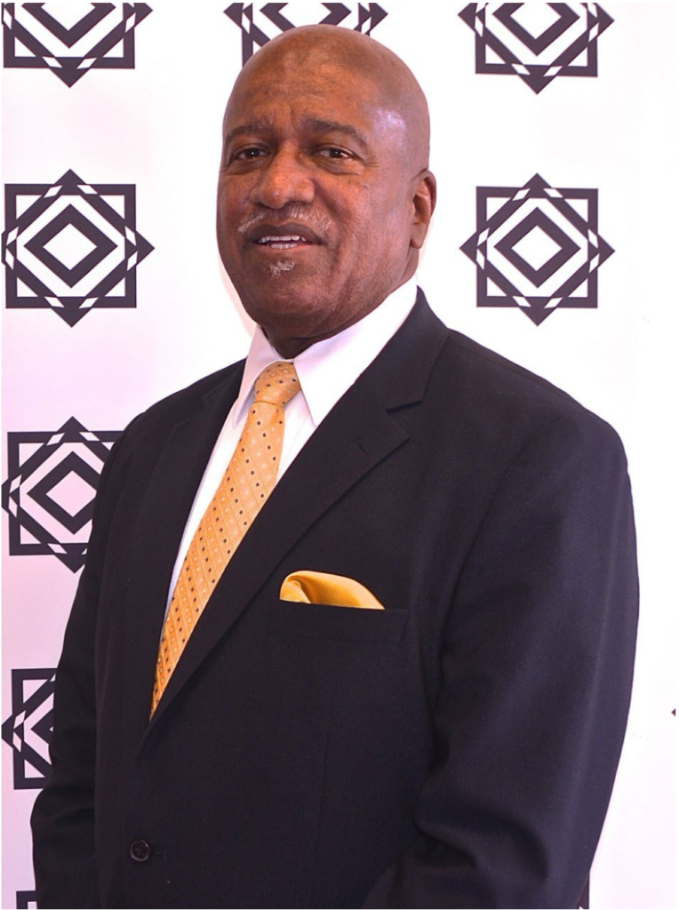 Arnold Benson in a dark suit with an orange tie and pocket square standing in front of a patterned backdrop, smiling at the camera.