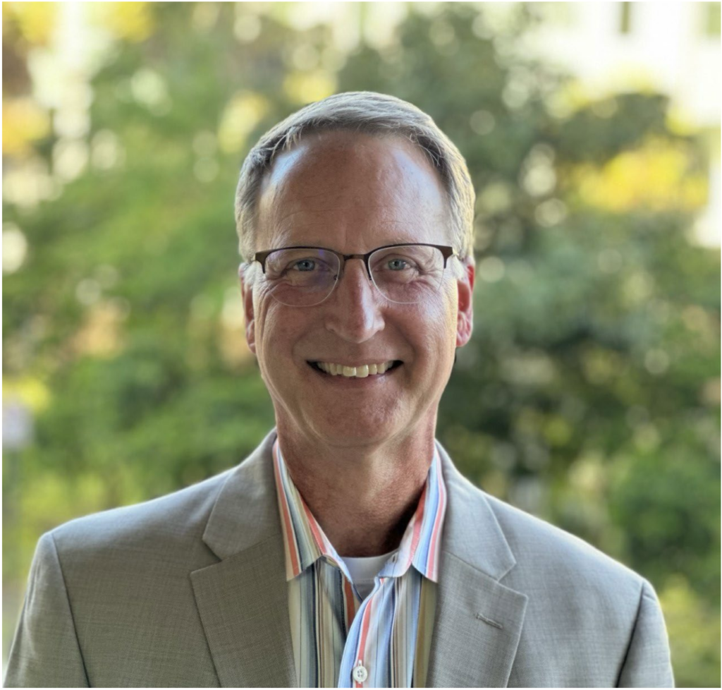 Portrait of Michael Fuhrman wearing glasses and a light-colored blazer, smiling outdoors with greenery in the background.