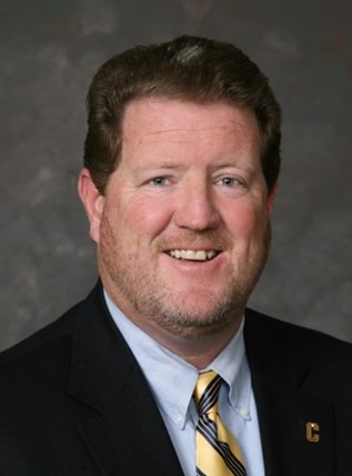 Headshot of Richard Cook, wearing a dark suit, light blue shirt, and striped tie, smiling against a neutral background.