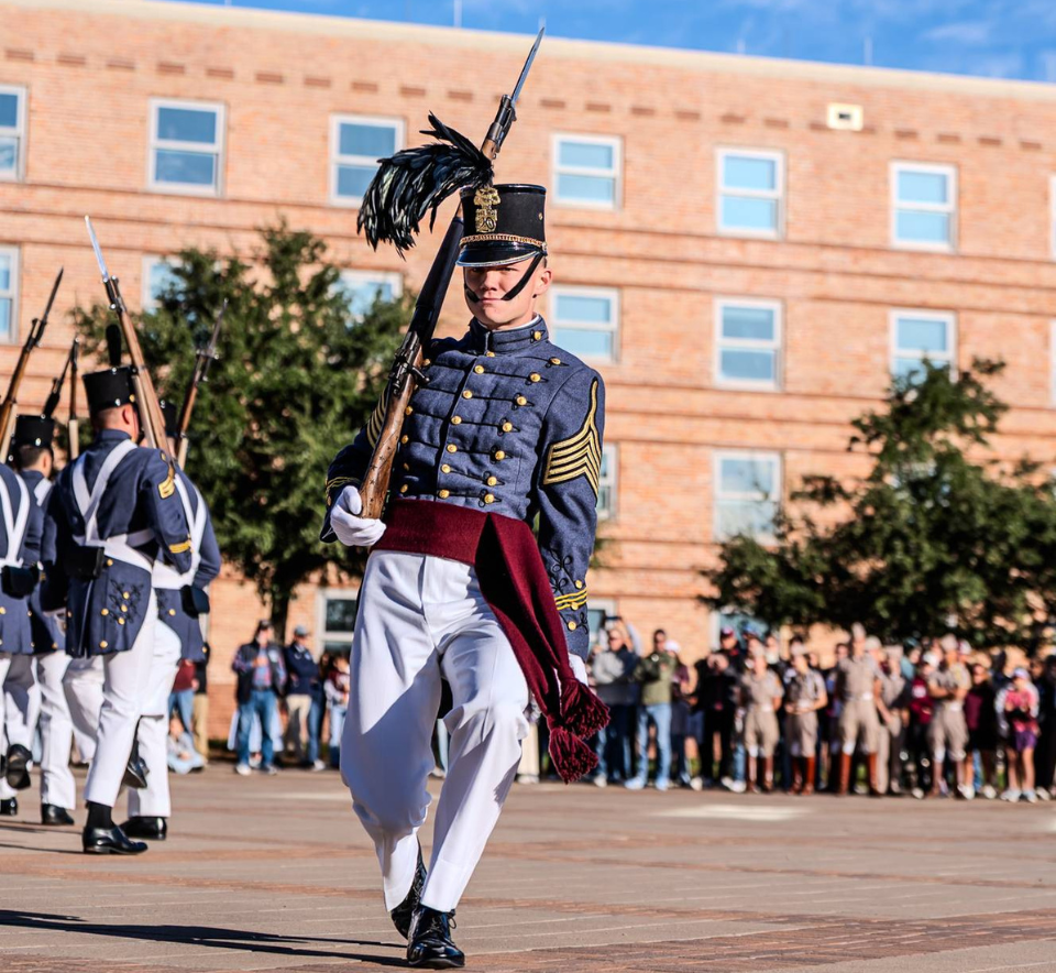 Citadel cadet in white dress uniform standing at attention during a parade, with fellow cadets carrying the South Carolina Corps of Cadets banner and flags behind him.