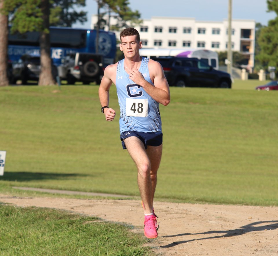 A runner wearing a light blue Citadel uniform with race number 48 runs on a dirt path during a track and field race.