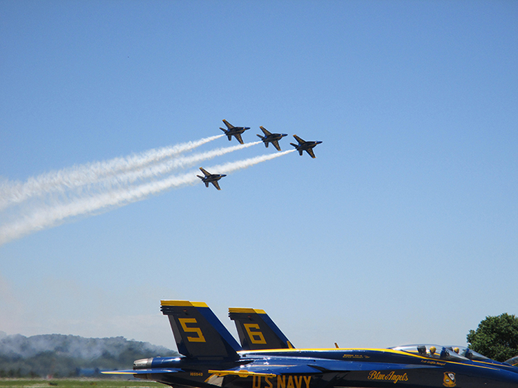 Photograph of grounded Blue Angels 5 and 6 with four unnamed Blue Angels flying above