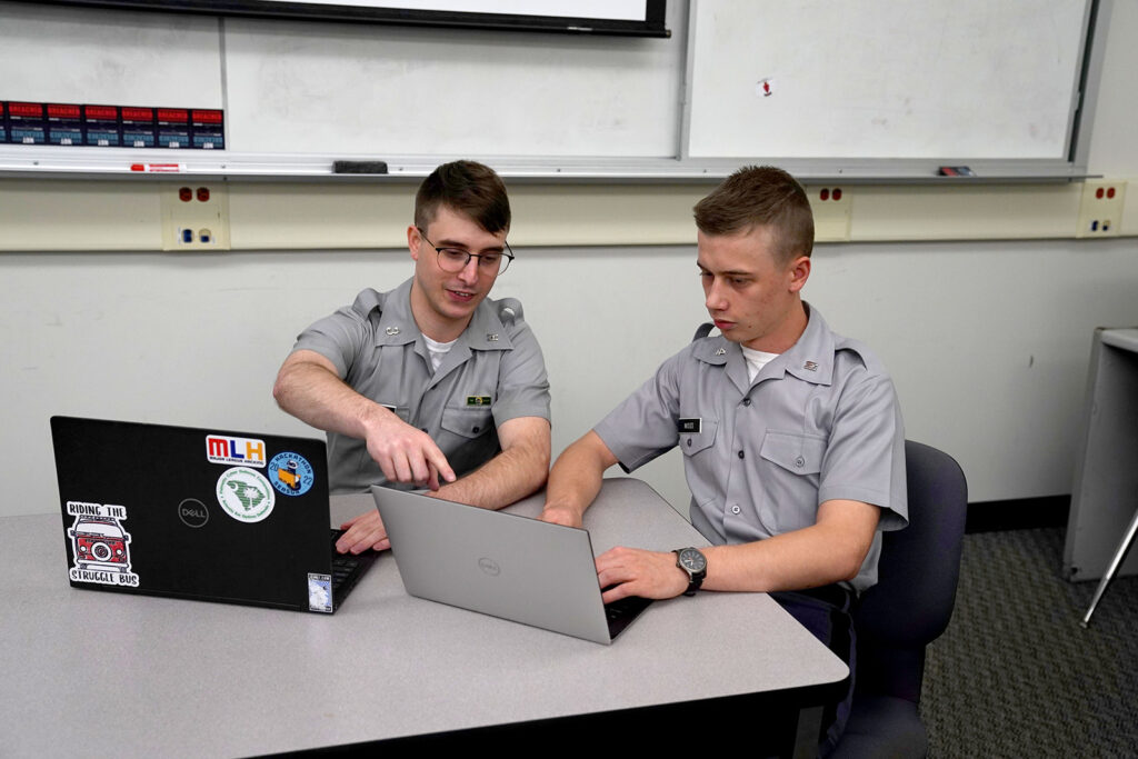 Two cadets collaborate on laptops at a table in a classroom.