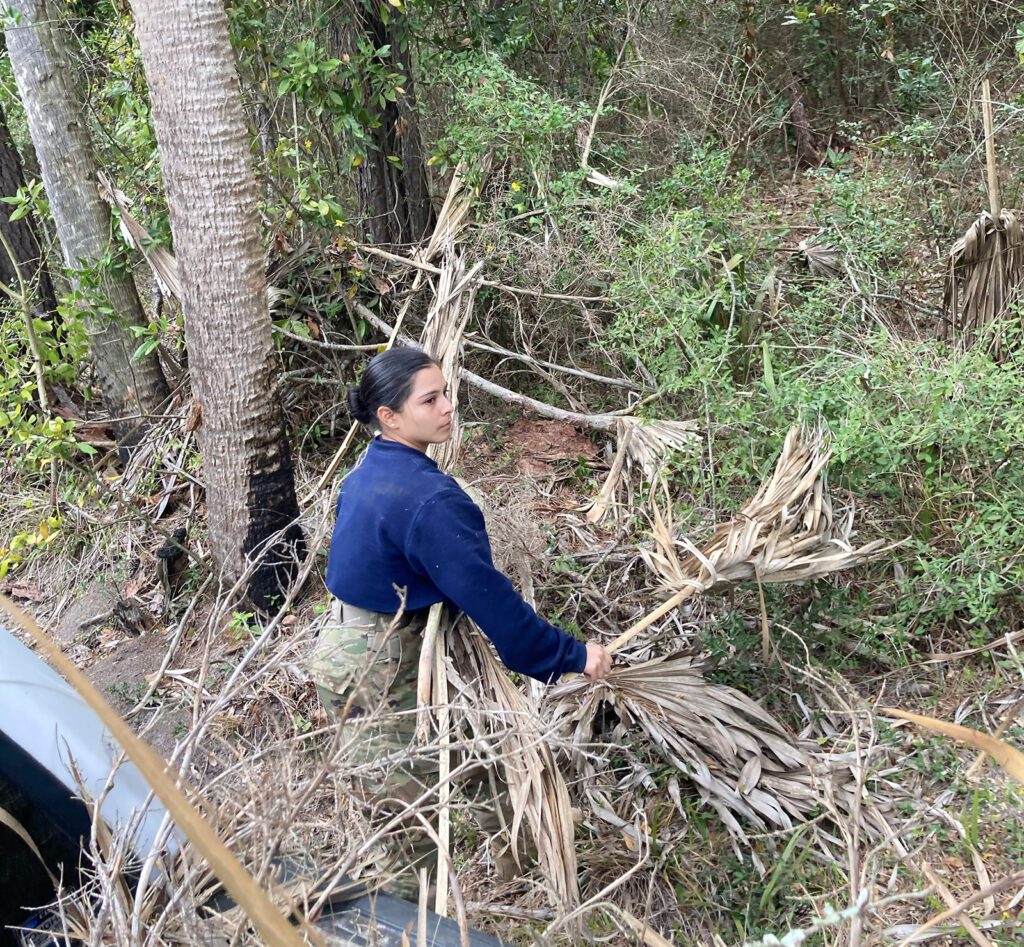Female cadet clears vegetation in a wooded area during an outdoor activity.