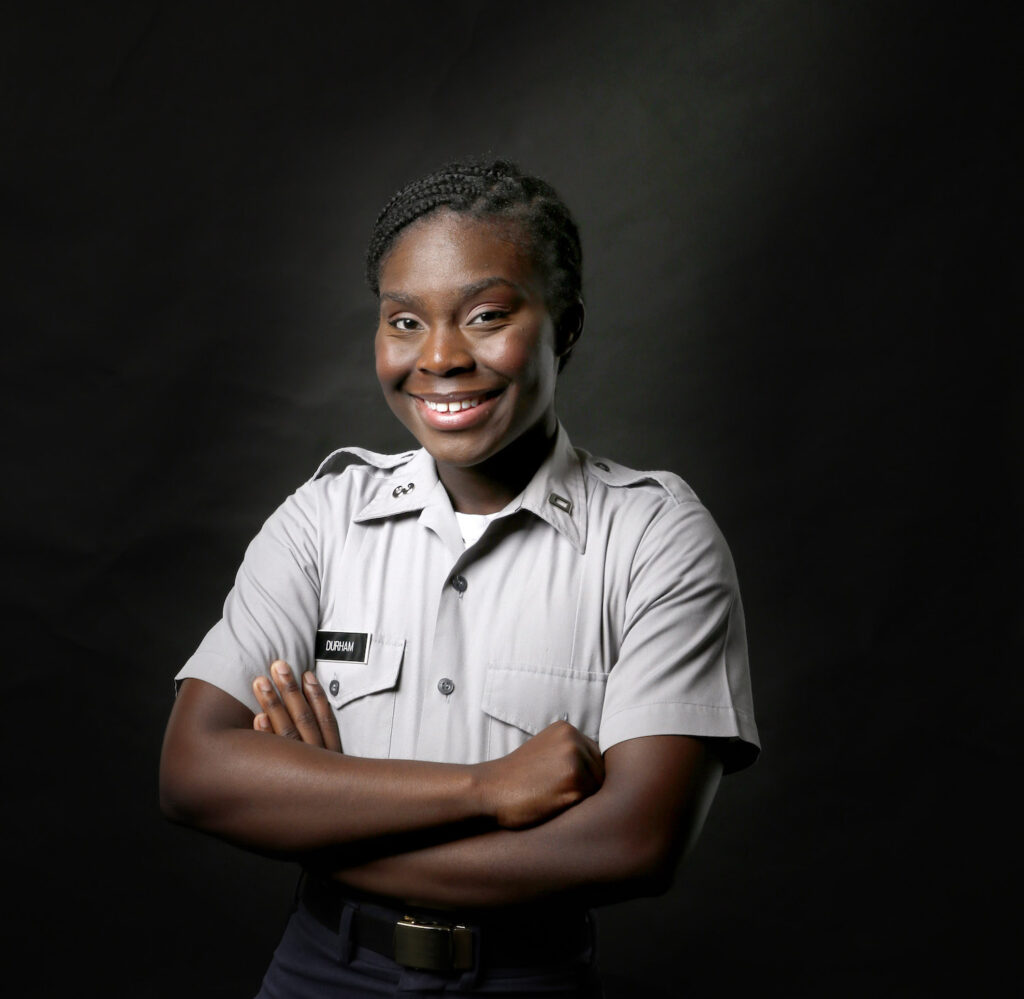 Smiling Citadel cadet Durham in a gray uniform with arms crossed against a black background.