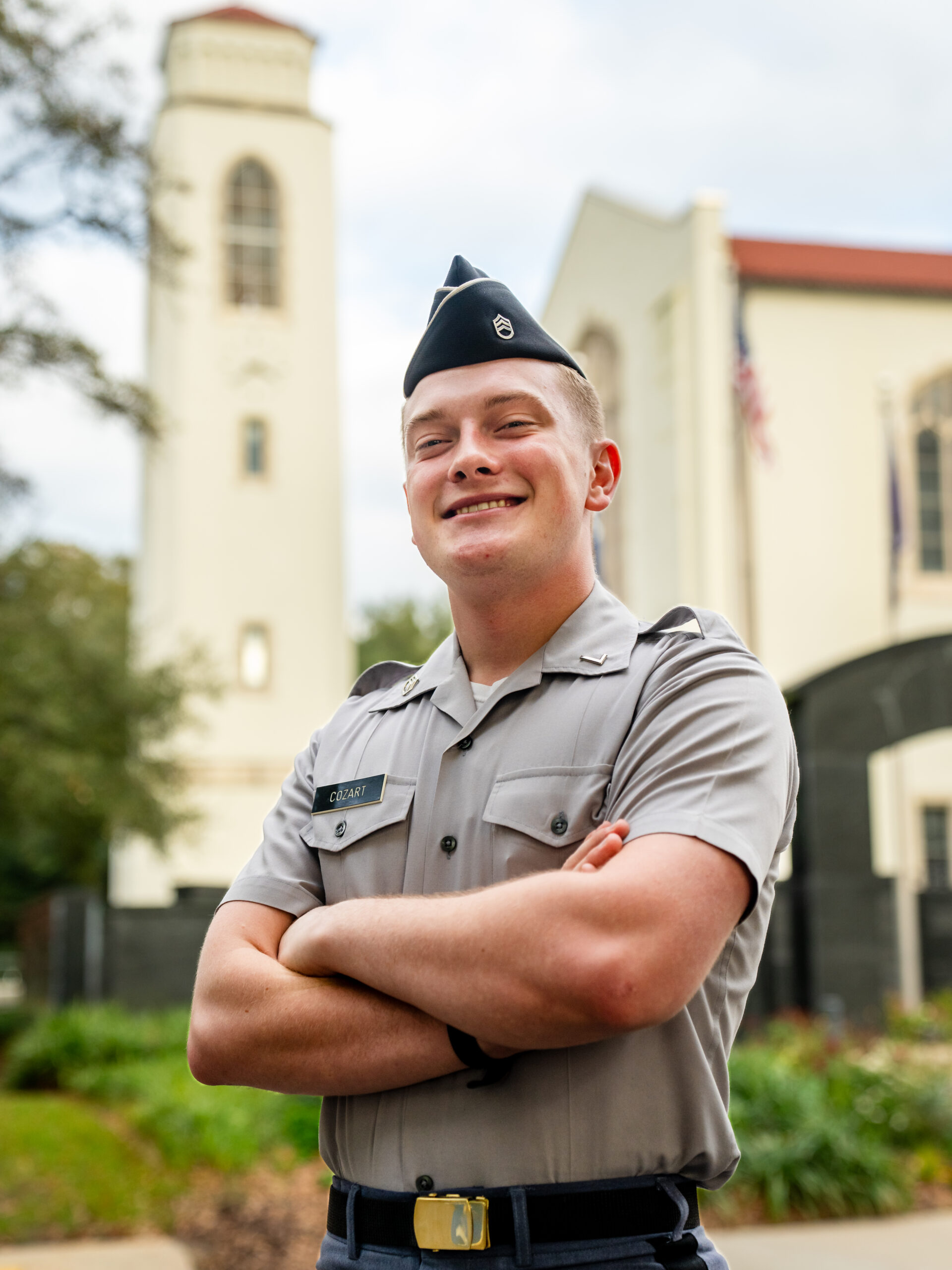 Cadet in gray uniform standing with arms crossed and smiling in front of Summerall Chapel on The Citadel campus.