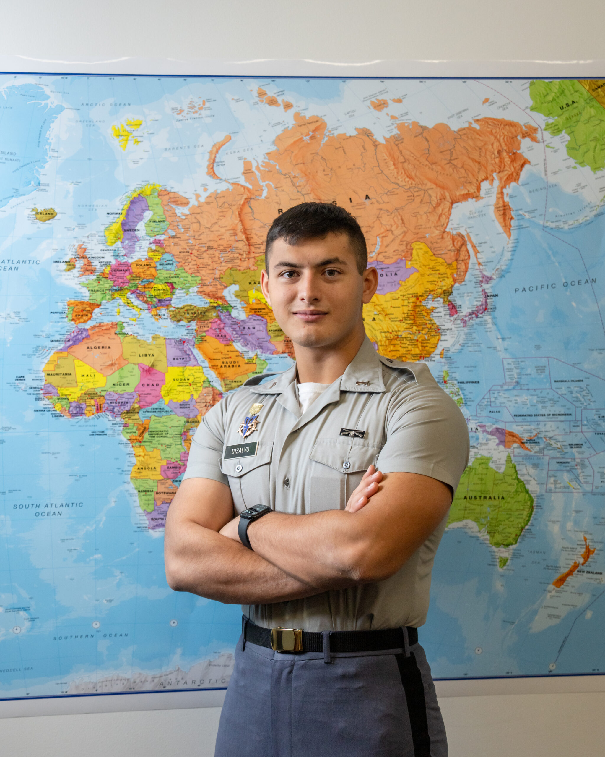 Cadet DiSalvo stands with arms crossed in military uniform in front of a large world map.