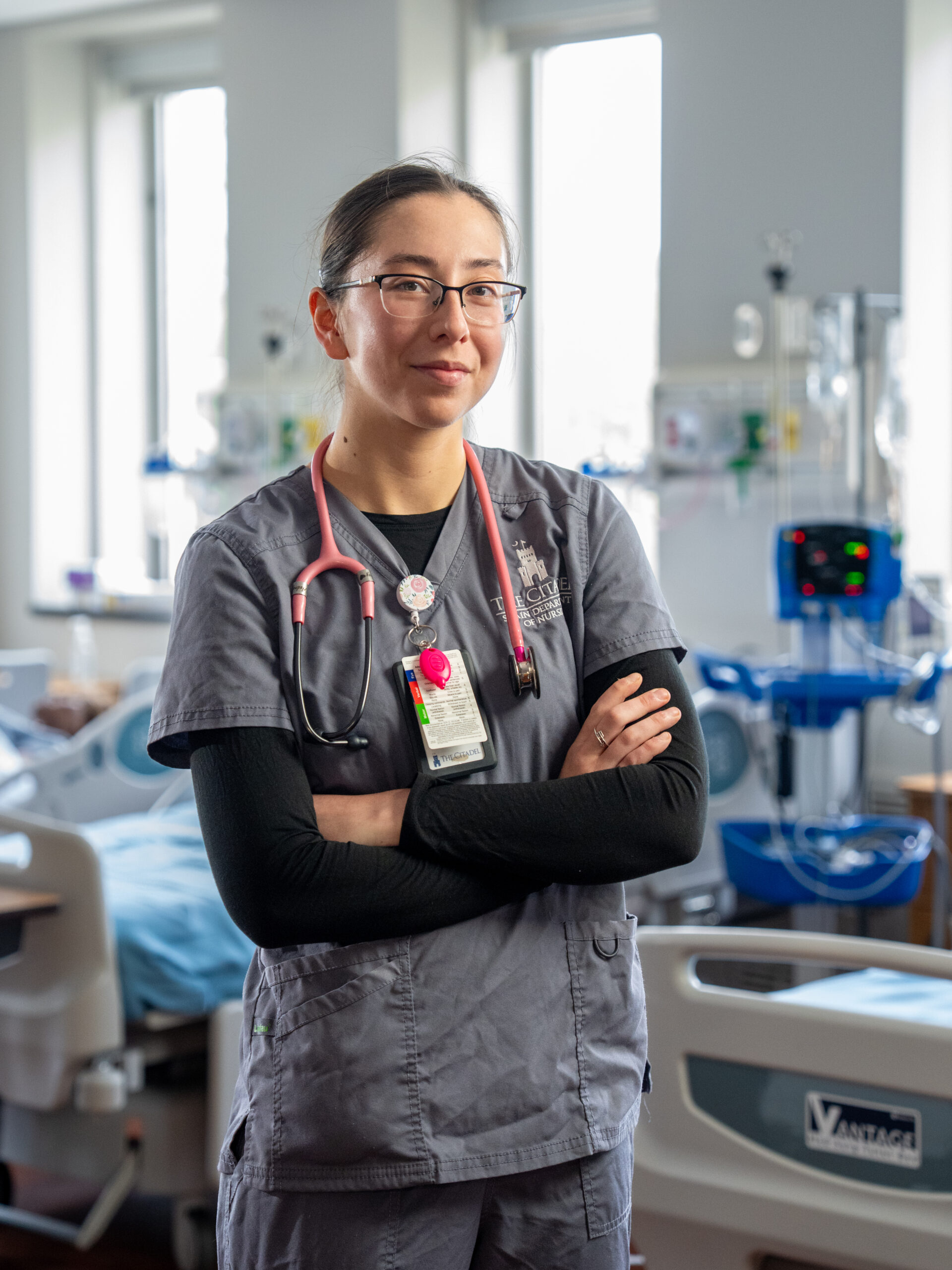 Nursing student in gray scrubs with a stethoscope stands in a clinical simulation lab with hospital beds and medical equipment in the background.