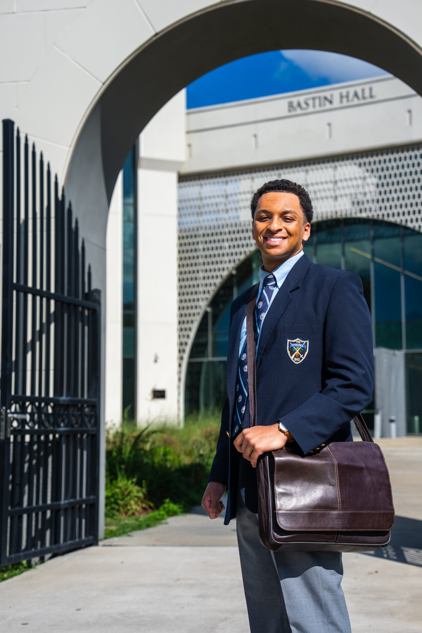 Cadet in dress uniform carrying a leather briefcase walks through an archway outside Bastin Hall, smiling toward the camera.