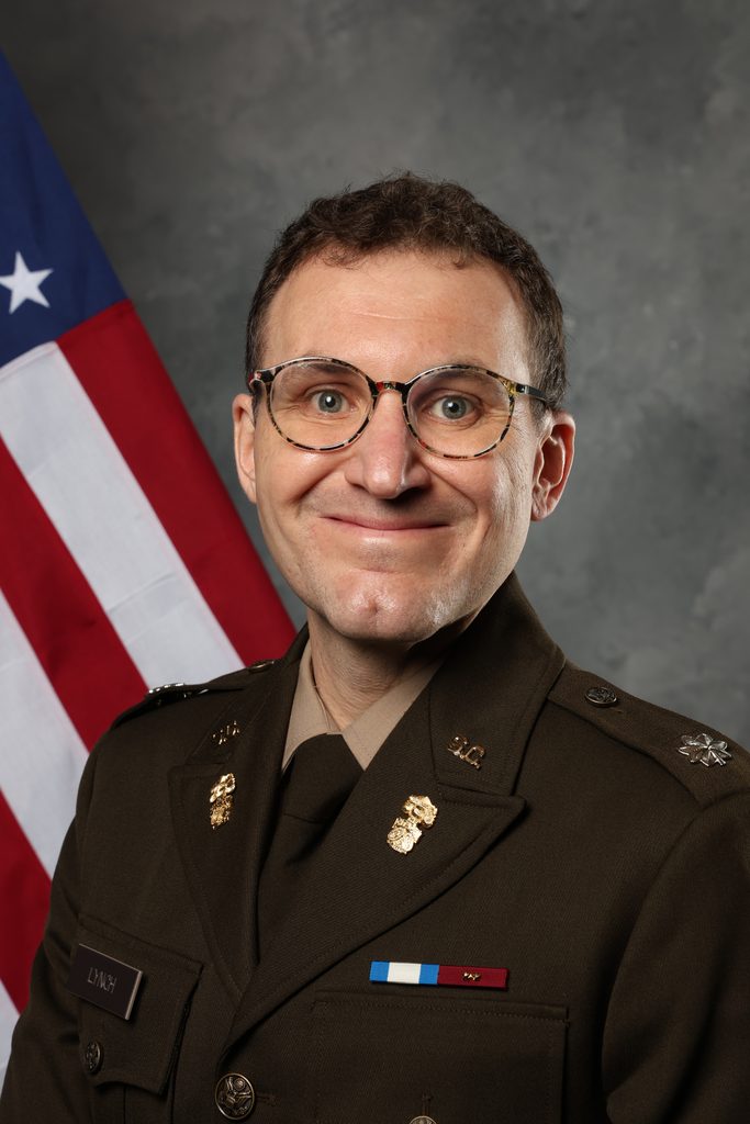 A portrait of Dr. Lynch in a military uniform with glasses, smiling in front of an American flag.