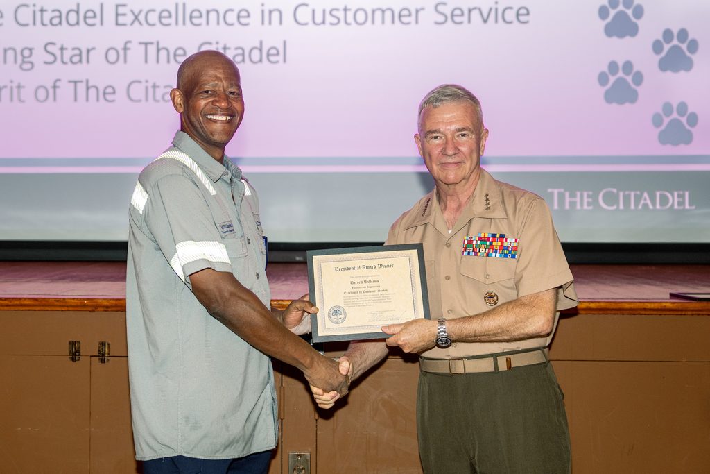 A person in a work uniform receives a framed certificate from another individual in a military-style uniform at an event hosted by The Citadel.