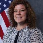 Angie Rochester with curly brown hair wearing a floral print top, posing in front of an American flag as part of the Citadel human resources team.
