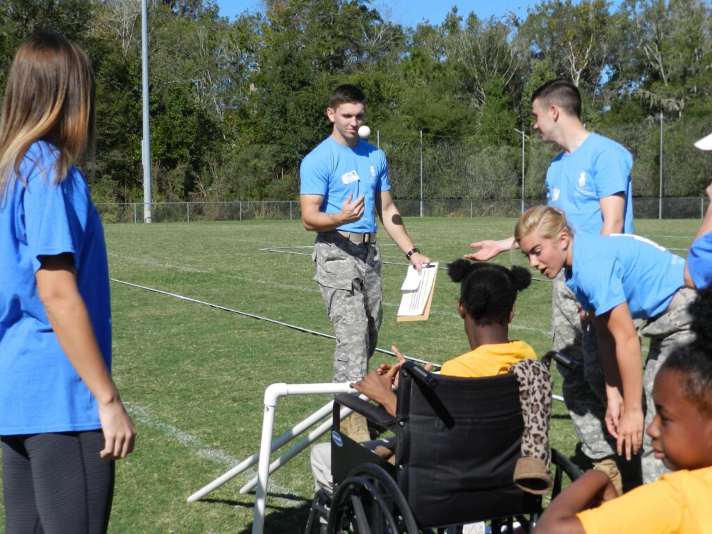 Citadel cadets in uniform participating in an outdoor activity with children on an athletic field.