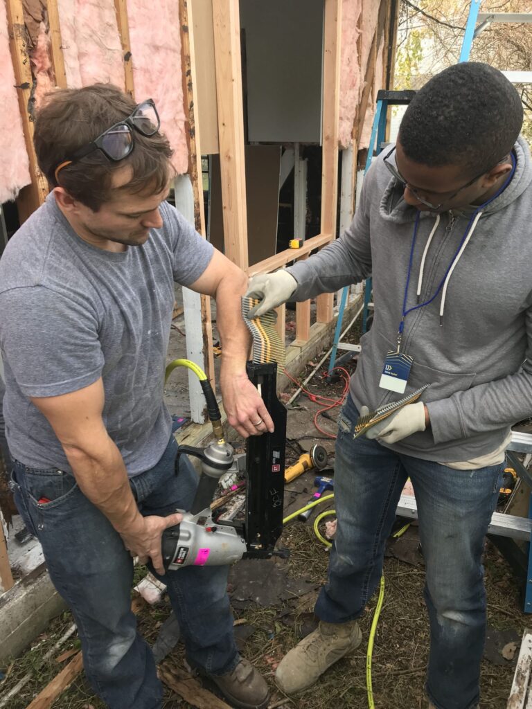 Two volunteers working with a pneumatic nail gun during a home construction or renovation project.