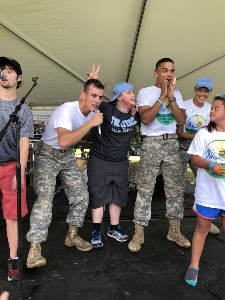 Citadel cadets and community participants gathered onstage during a Buddy Walk event, with one cadet speaking into a microphone.