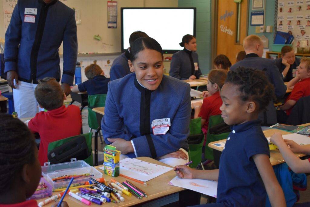 Citadel cadet working with an elementary school student on an art activity in a classroom while other cadets and students work nearby.