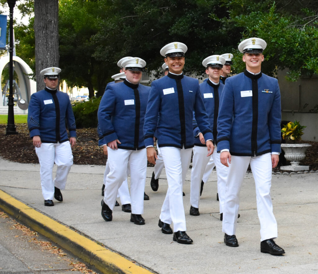 Citadel cadets in dress uniforms walk together outdoors on campus.