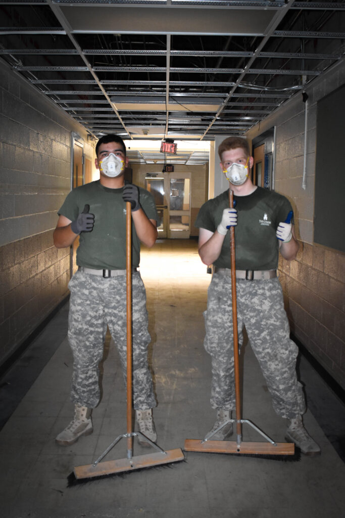 Two Citadel cadets wearing protective masks and gloves hold brooms while working inside a building during a service project.