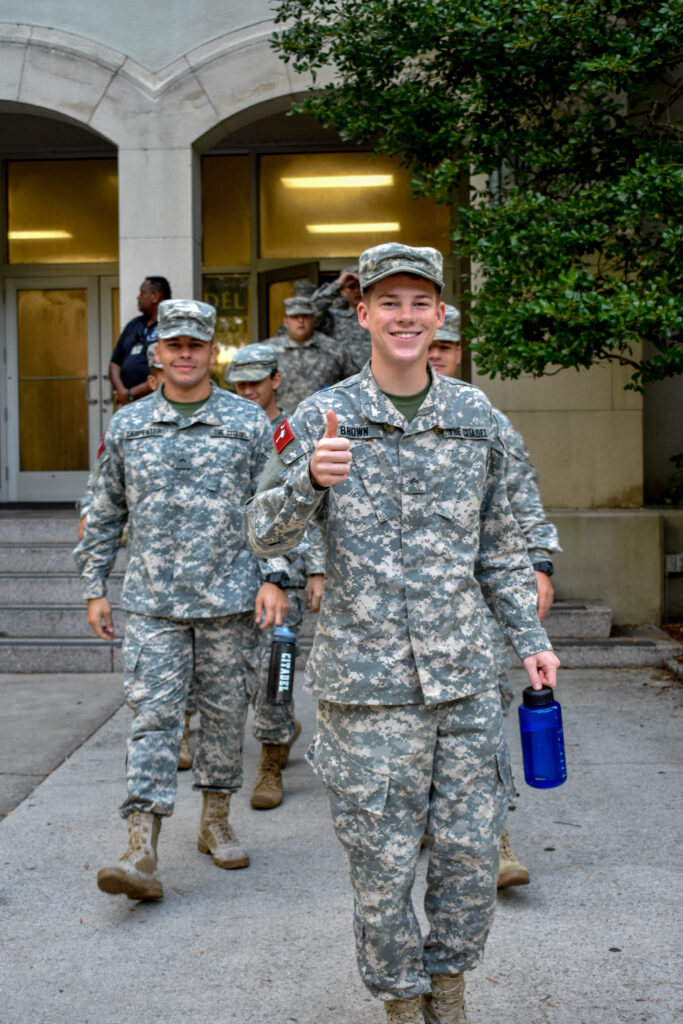 Citadel cadet in camouflage uniform gives a thumbs-up while walking with fellow cadets.