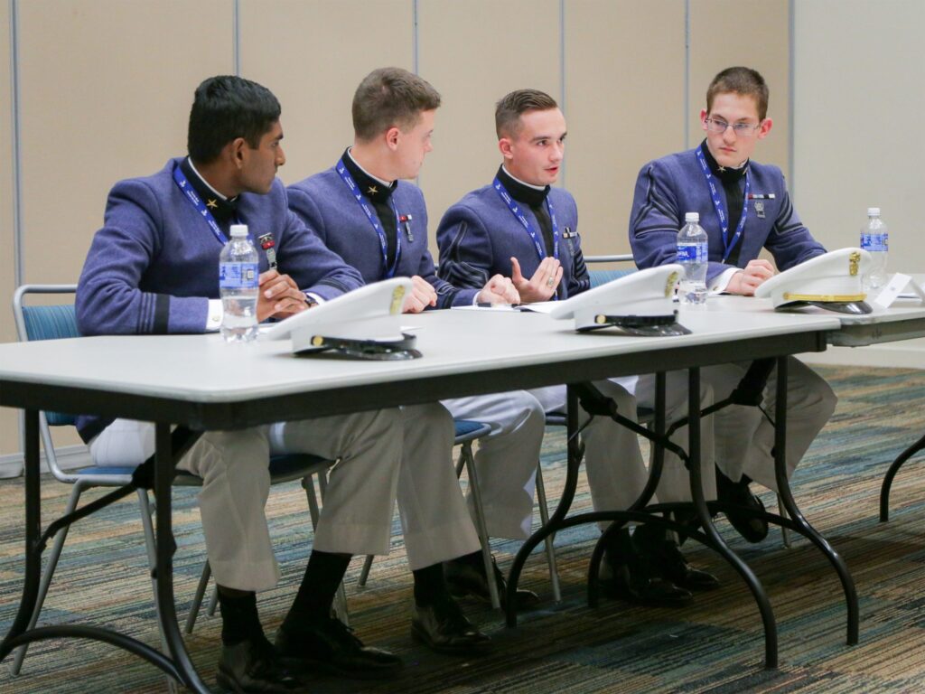 Four Citadel cadets in dress uniforms participate in a panel discussion, with one cadet speaking to the group.