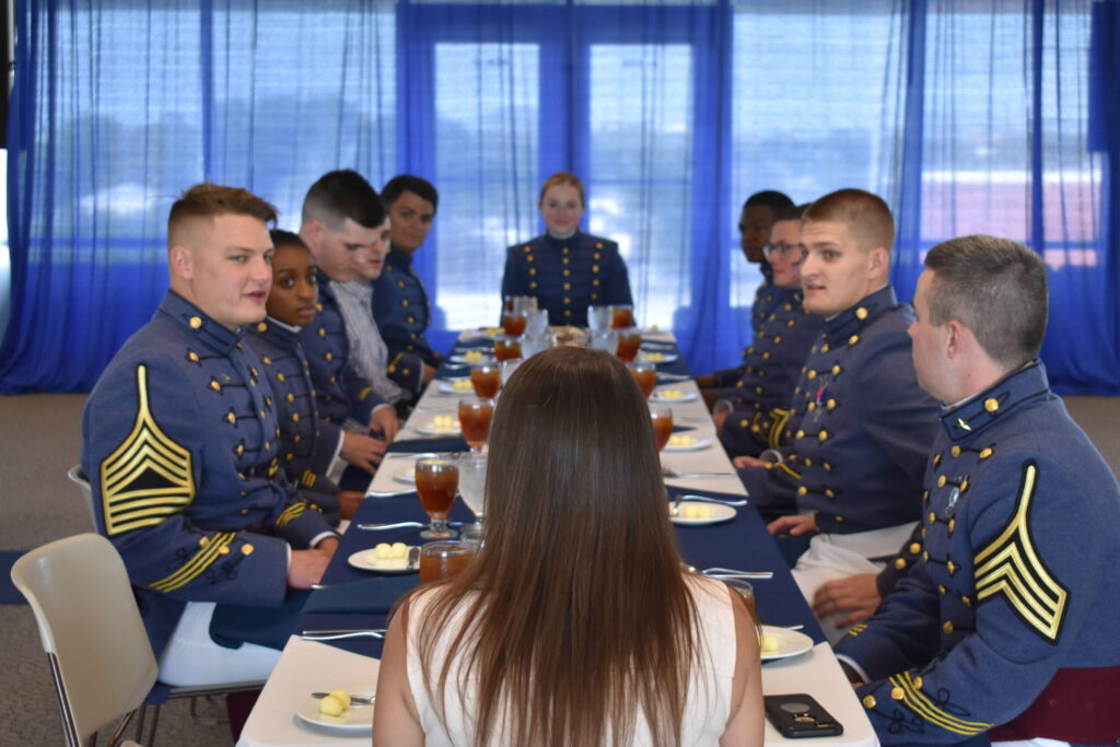Citadel cadets in dress uniforms participate in an etiquette training session around a formal dining table.