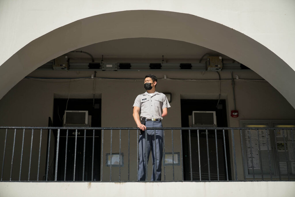 Cadet in uniform and a face mask standing on a barracks balcony beneath a large arch.