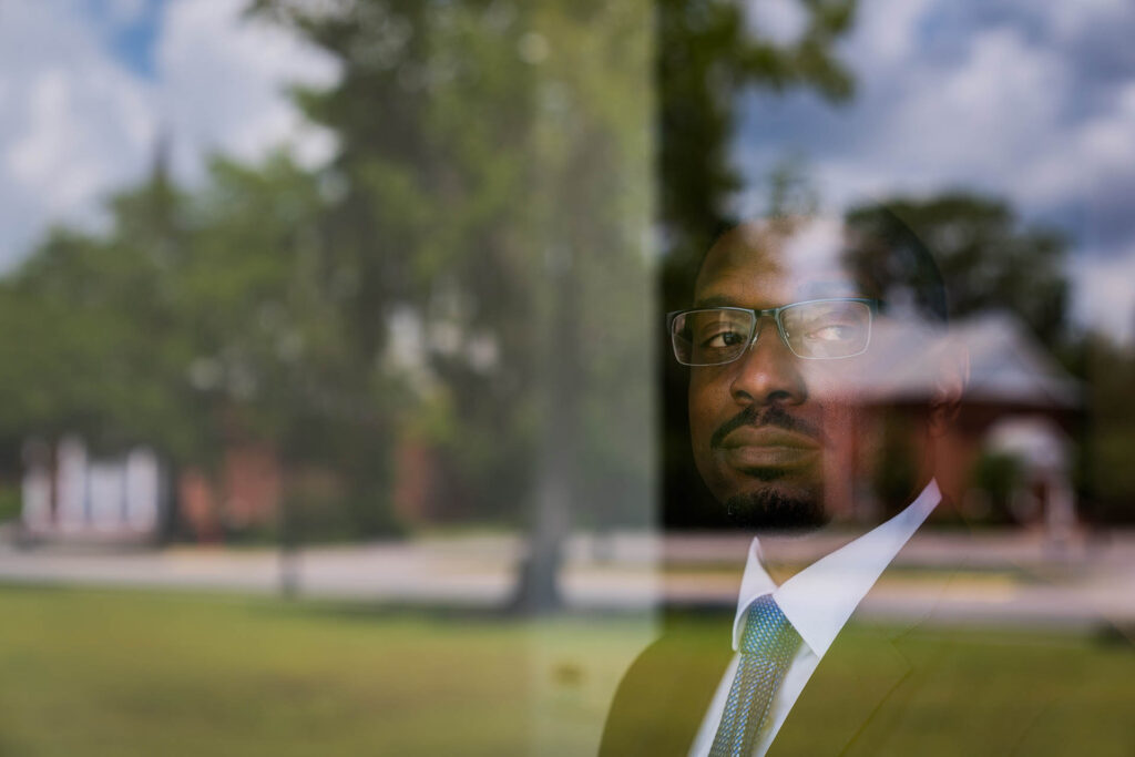 Man in a suit and glasses seen through a reflective window overlooking campus.